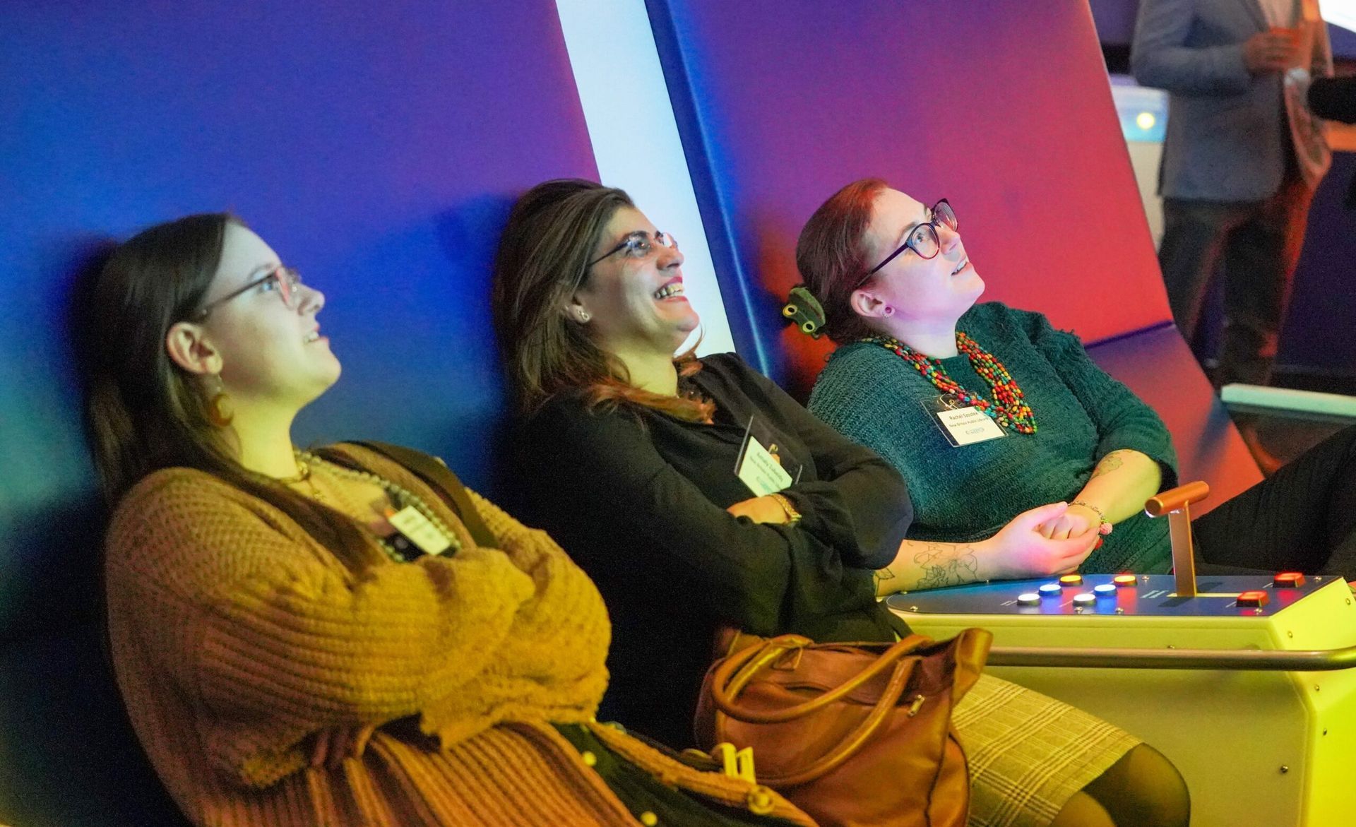 Three people looking up and laughing; seated, leaning against blue and white walls with colorful lighting.
