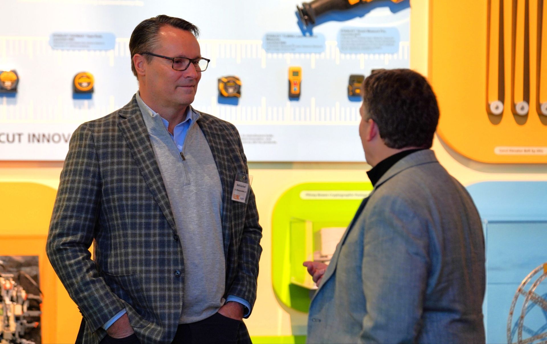 Two men talking; one in plaid jacket, the other in a gray coat, near a colorful wall with equipment displays.