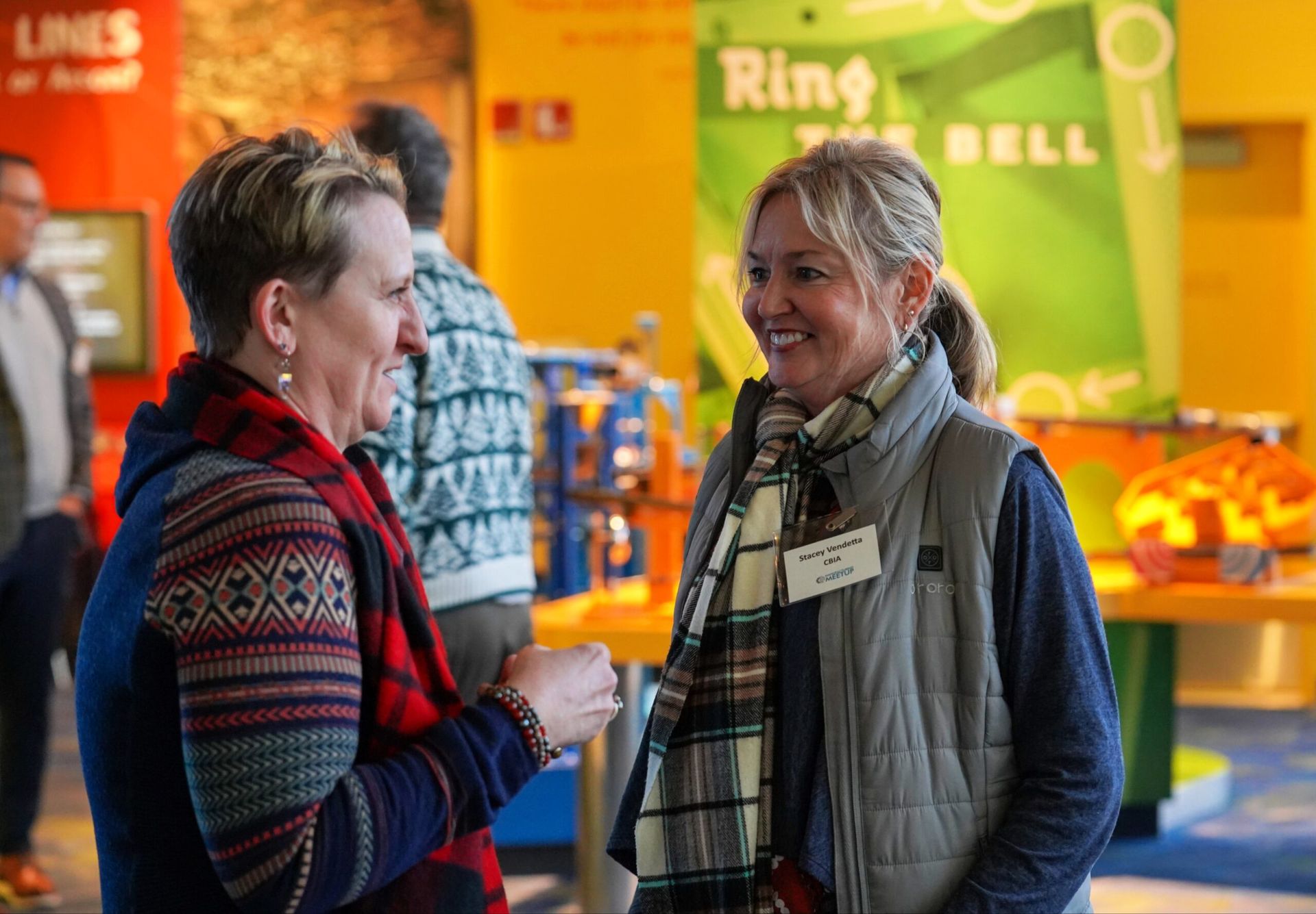 Two women chatting at an event. One wears a blue sweater, patterned scarf; the other, a vest, scarf, and name tag. Bright background.