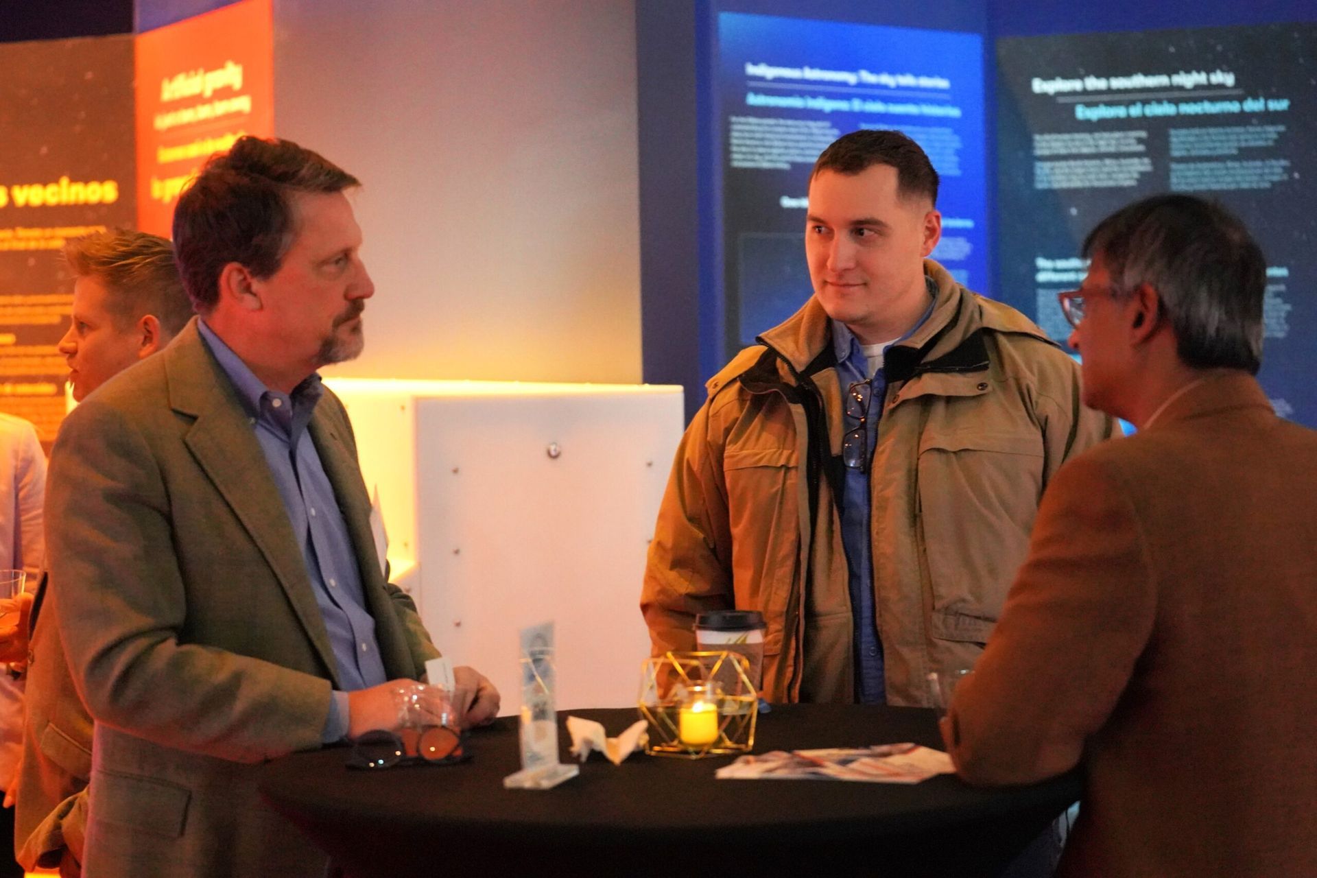 Three men in conversation at an event, standing near a small table.
