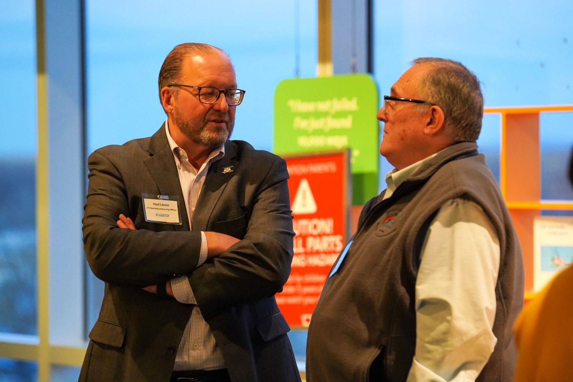 Two men in conversation, one in a suit, the other in a vest, standing indoors near informational signs.