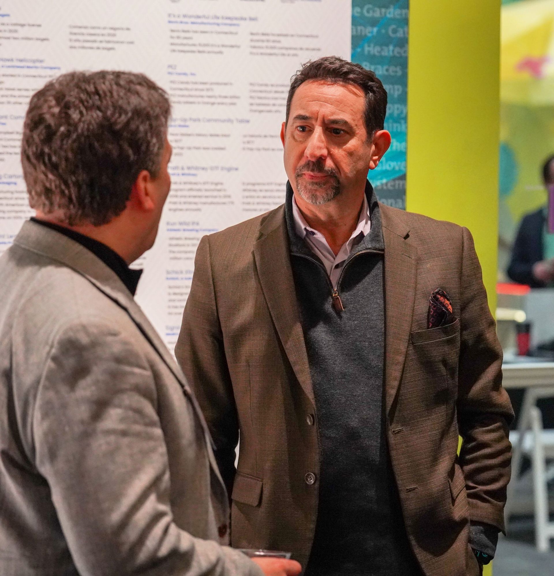 Two men in business attire conversing at a conference; one in a brown blazer, the other in a light suit.