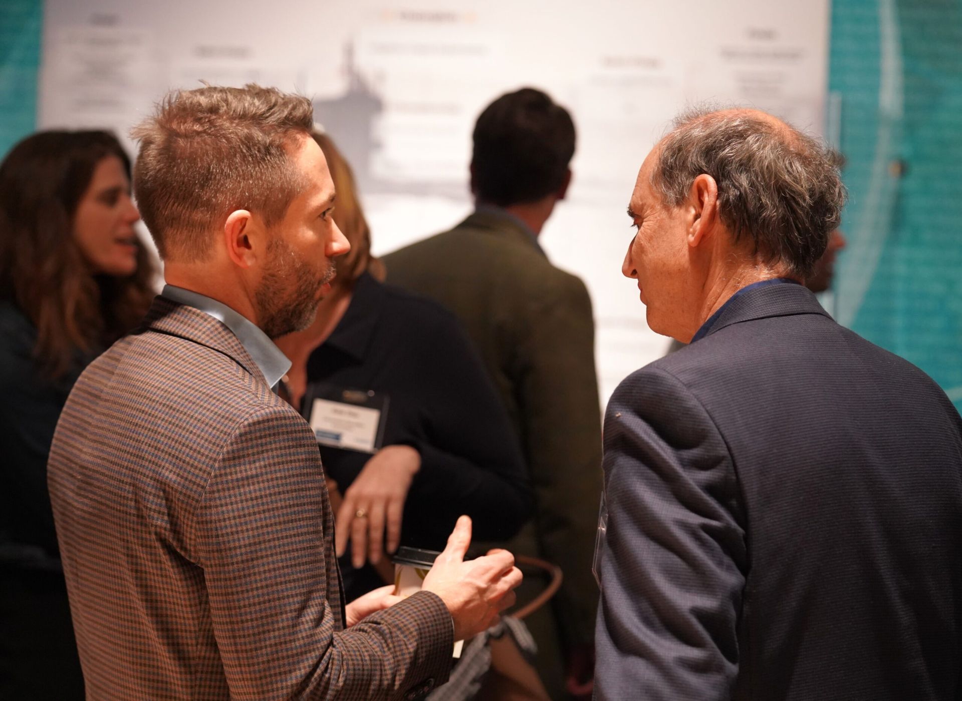 Two men converse at an event, standing near a poster with others in the background.