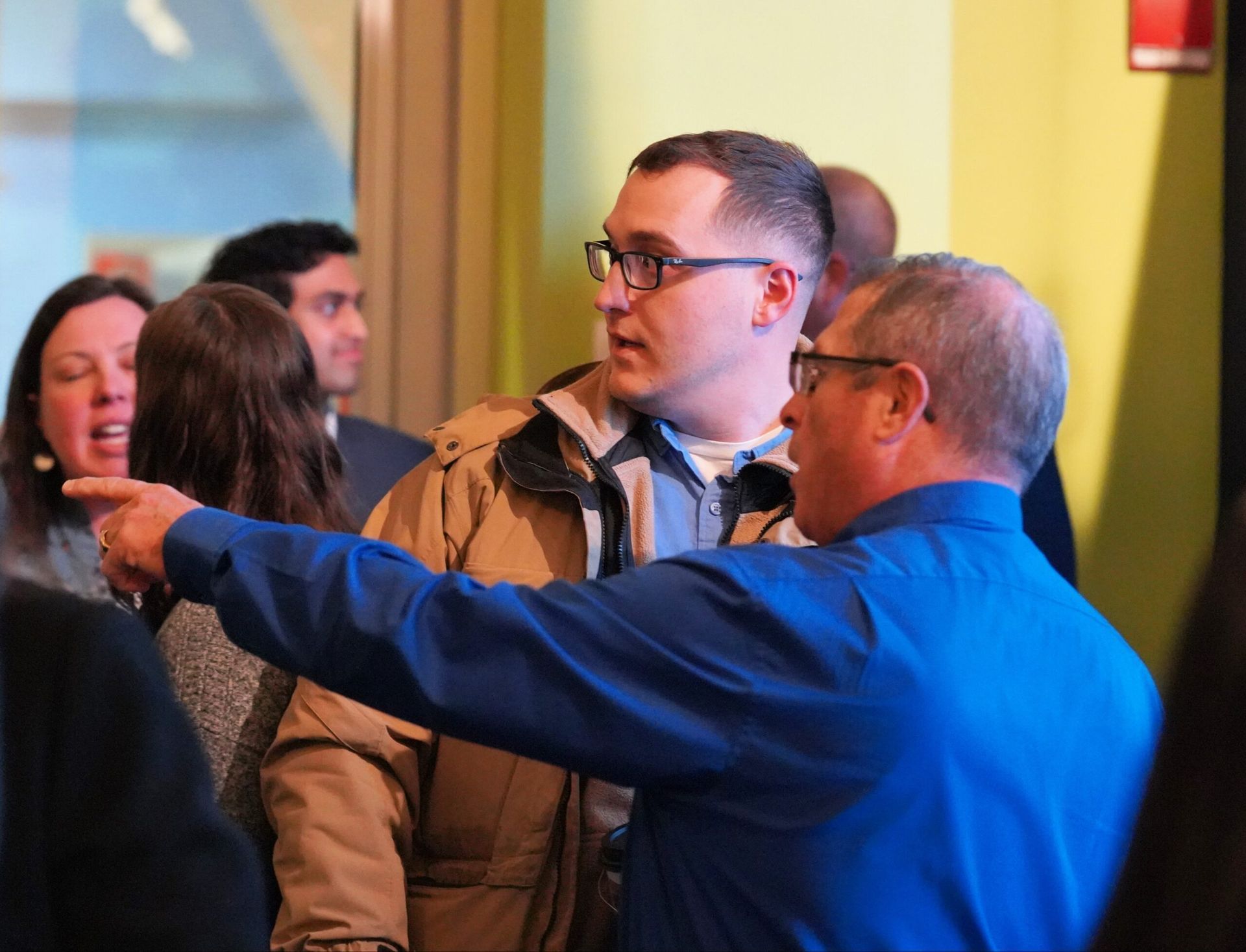 Man in tan coat and glasses conversing with man in blue shirt, pointing to the left. People in background.
