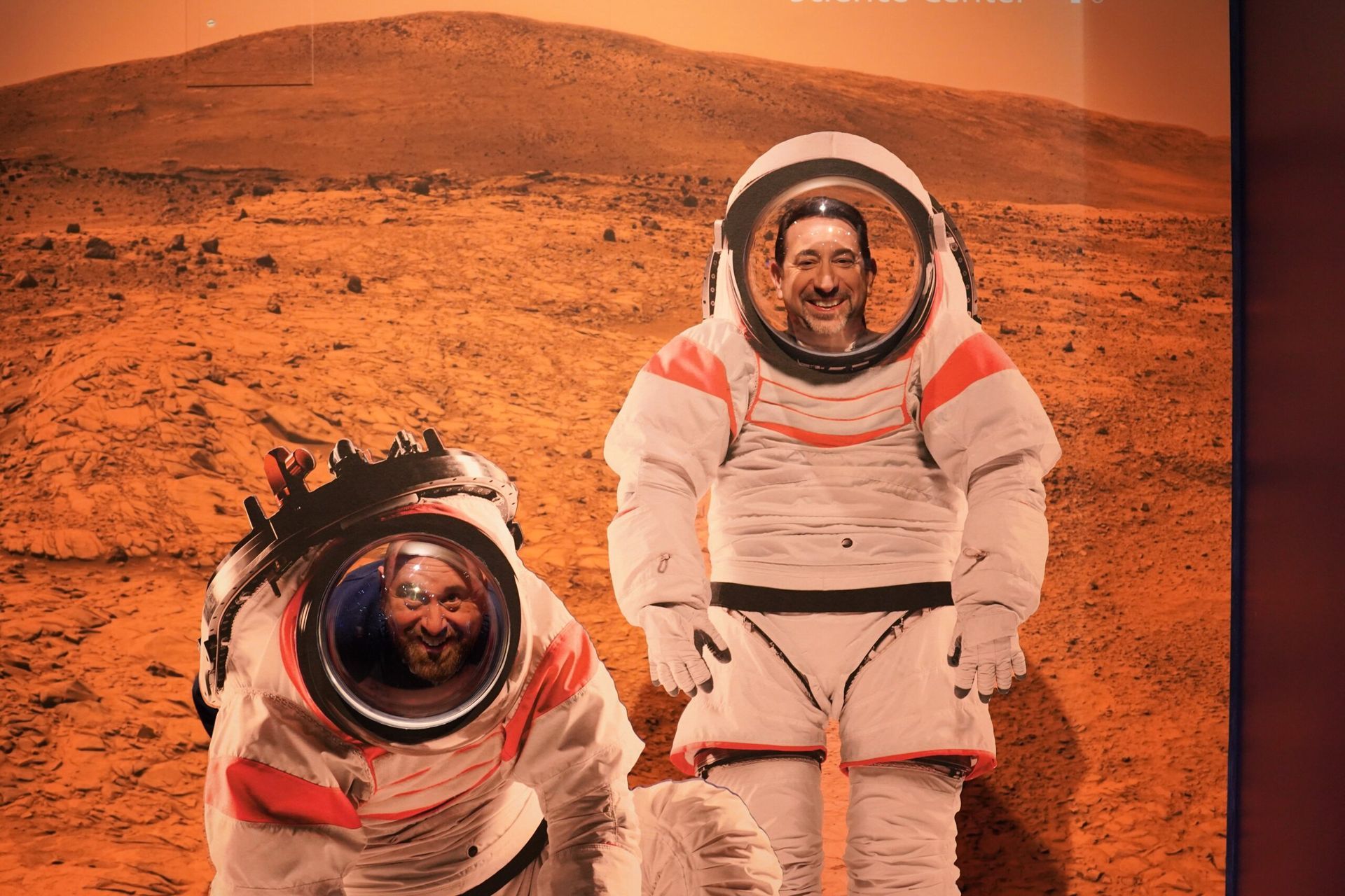 Two people in astronaut suits posing in front of a Mars backdrop. One stands, the other kneels, both smiling.