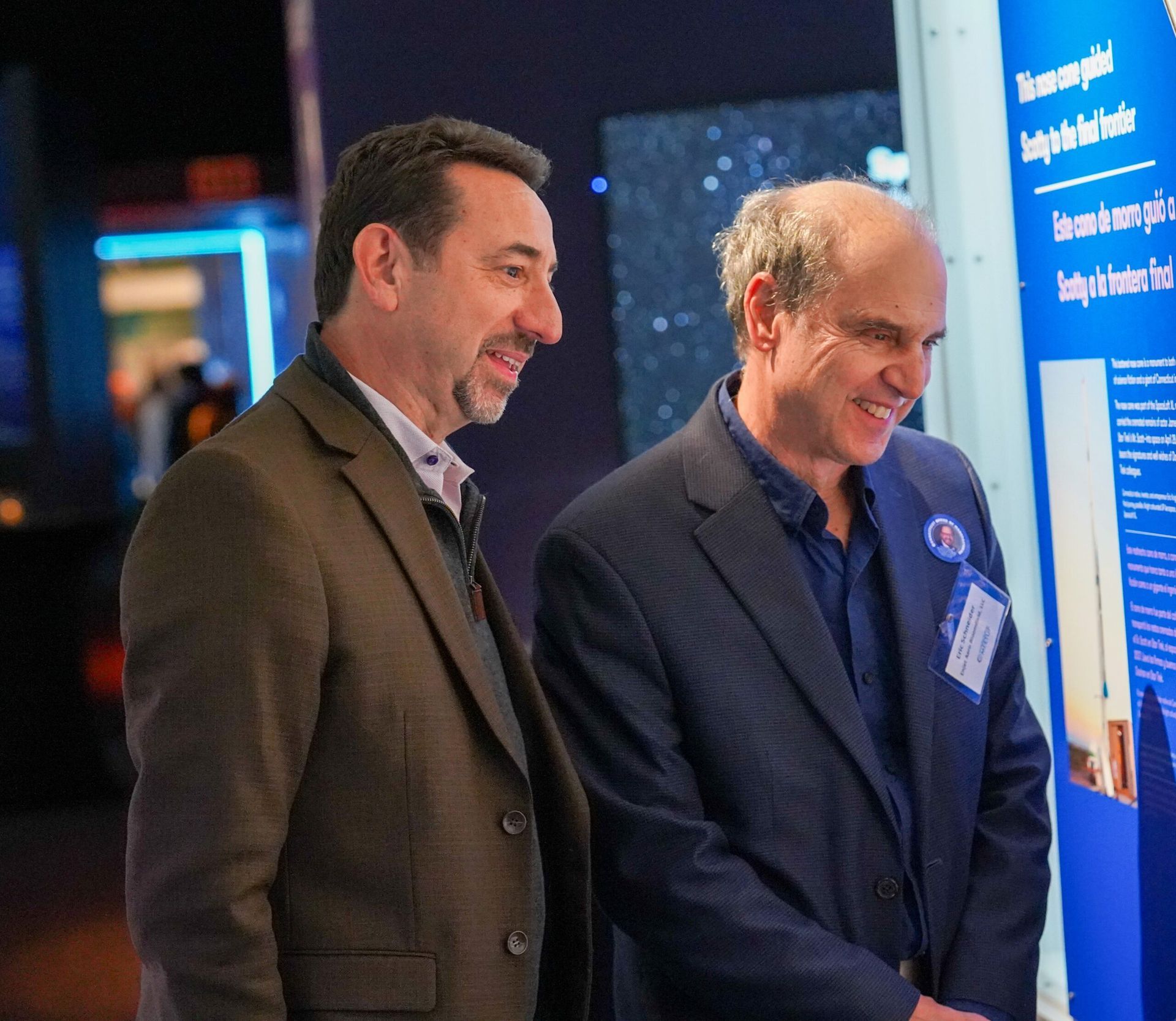 Two men looking at a museum display, smiling. One wears a brown jacket, the other a blue blazer.