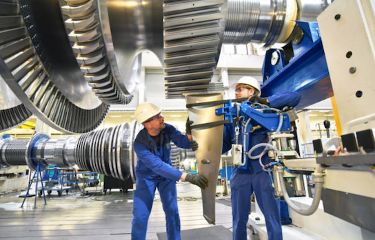 Two engineers in blue coveralls and hard hats working on large machinery in a factory.