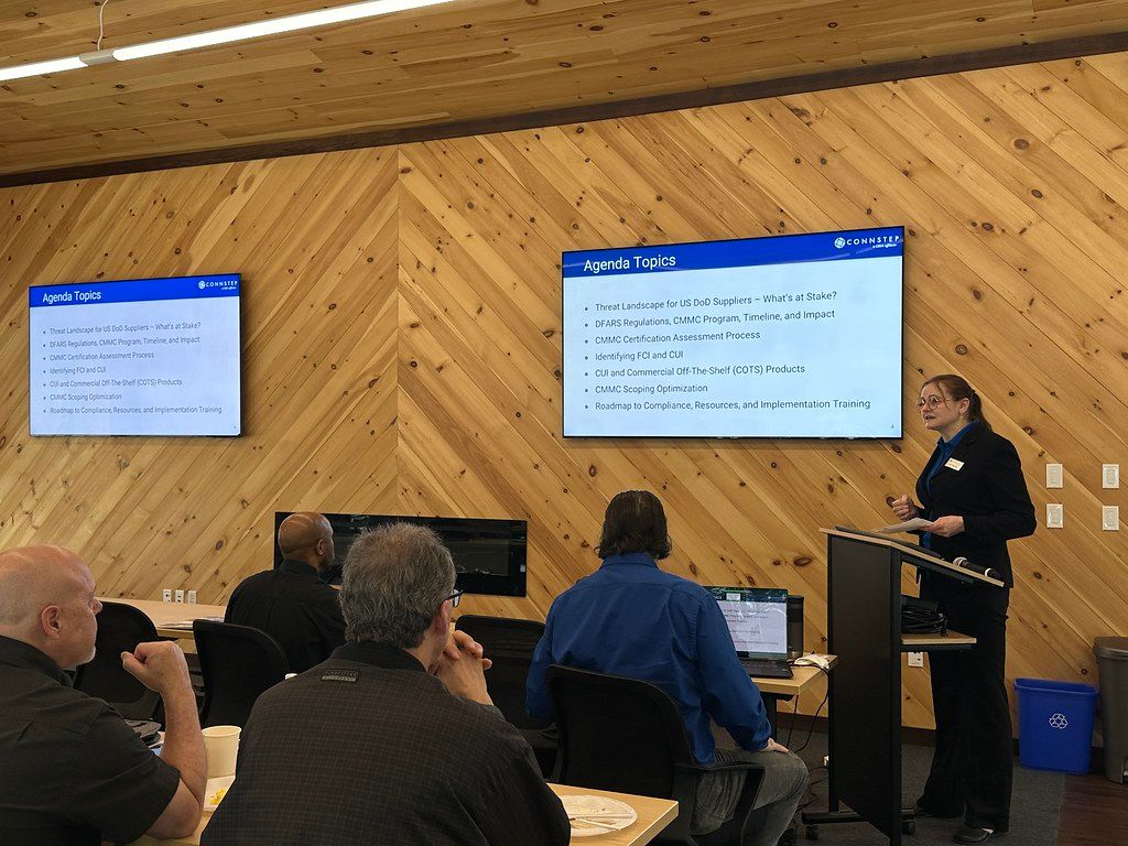 A presenter at a conference giving a presentation to a seated audience with screens displaying text.