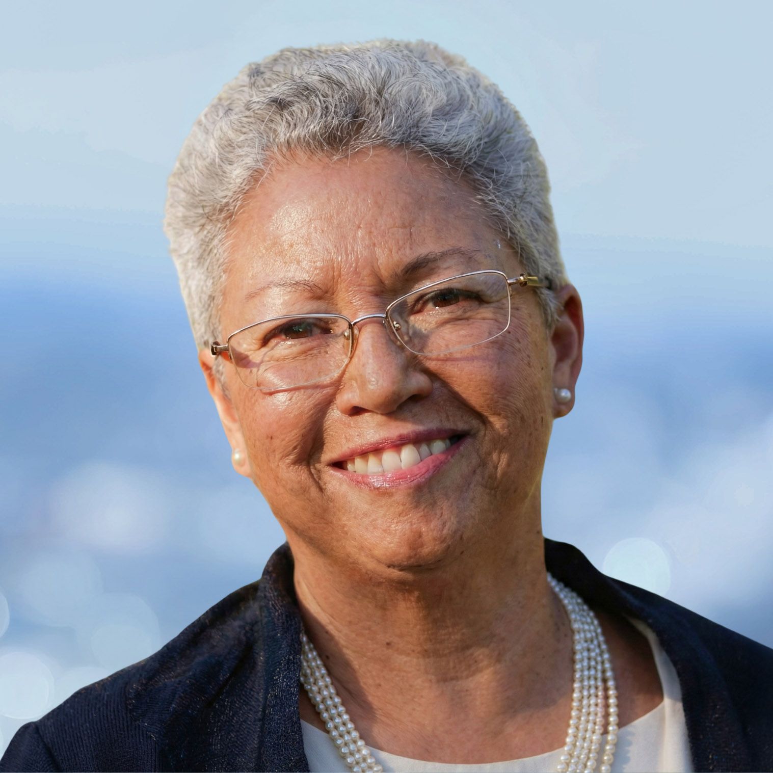 Woman with gray hair and glasses smiling, wearing pearls and a navy jacket.