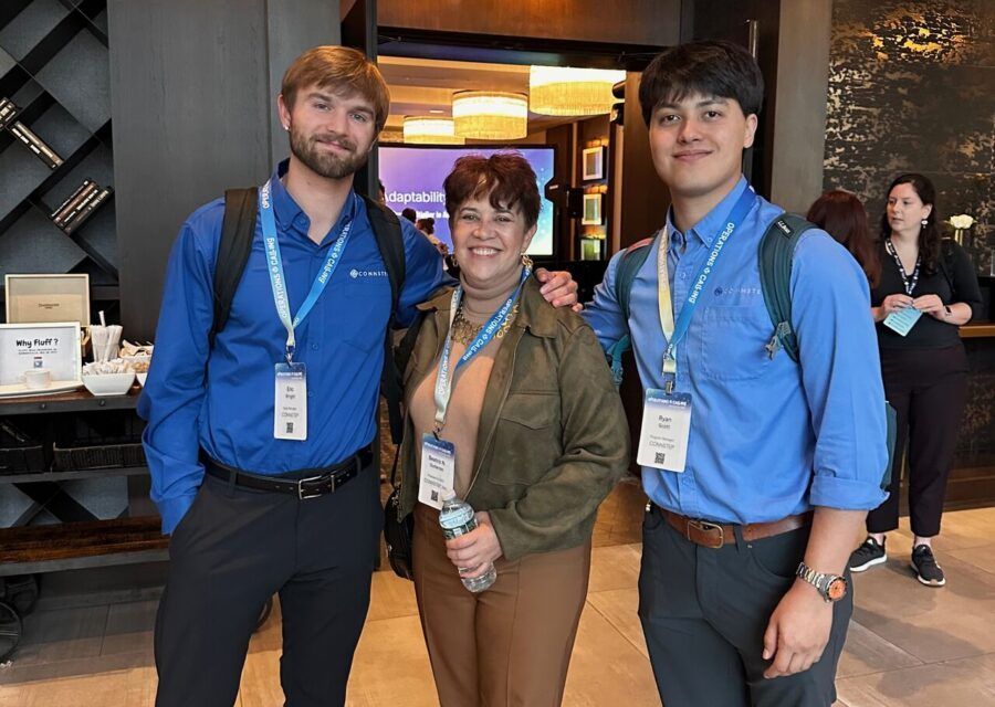 Three people, two in blue shirts and one in a brown jacket, stand together smiling indoors.