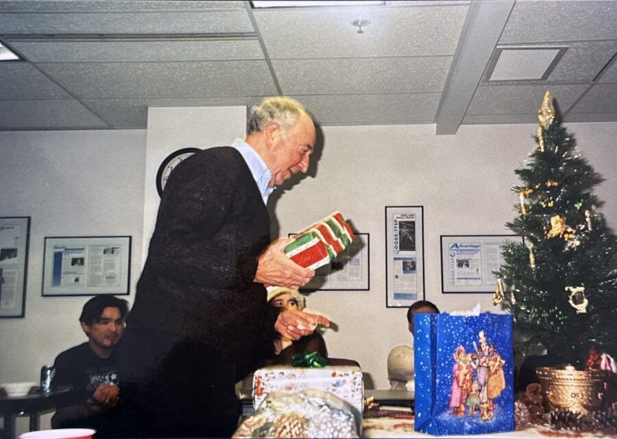 Man opening a Christmas gift near a small tree. A younger person watches. Office setting.