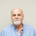 Man with a white goatee wearing a blue button-down shirt.  Against a plain, off-white background.