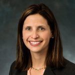 Woman with dark hair smiles, wearing a black blazer and light-colored top; neutral background.