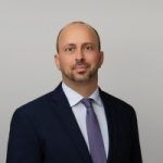 Man with short brown hair, wearing a navy suit, white shirt, and purple tie, posing.