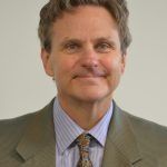 Man in a suit smiles, indoors; neutral background.