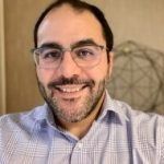Man with glasses smiling, light blue collared shirt, and short dark beard.