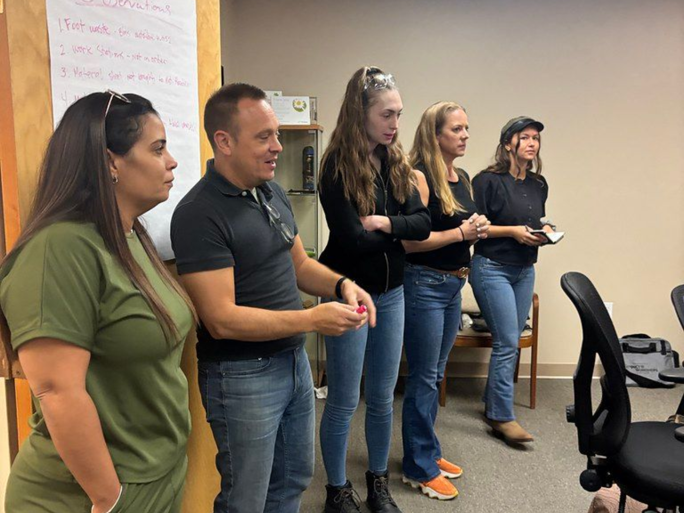 Five people stand in a room, listening. Two people wear jeans, a woman in a green outfit, another in a black shirt and cap.