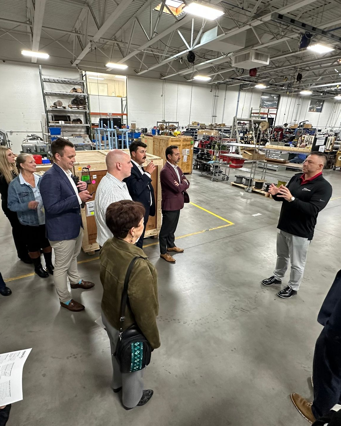 Group listens to a man giving a presentation in a warehouse. People stand in a semi-circle.