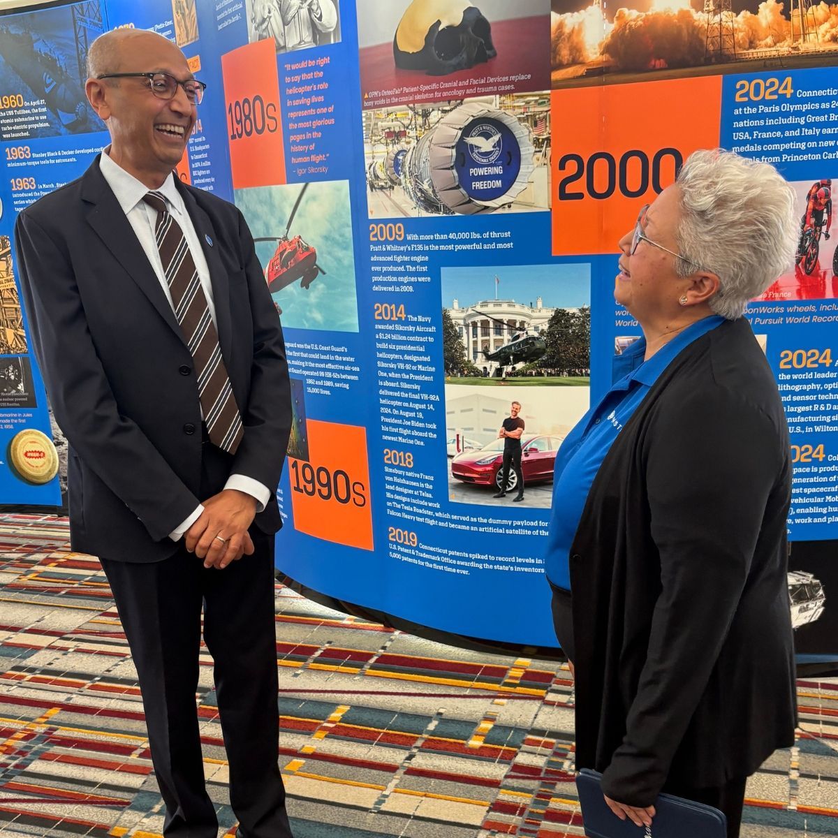 Man in suit laughs with woman in blue shirt, in front of a timeline display.