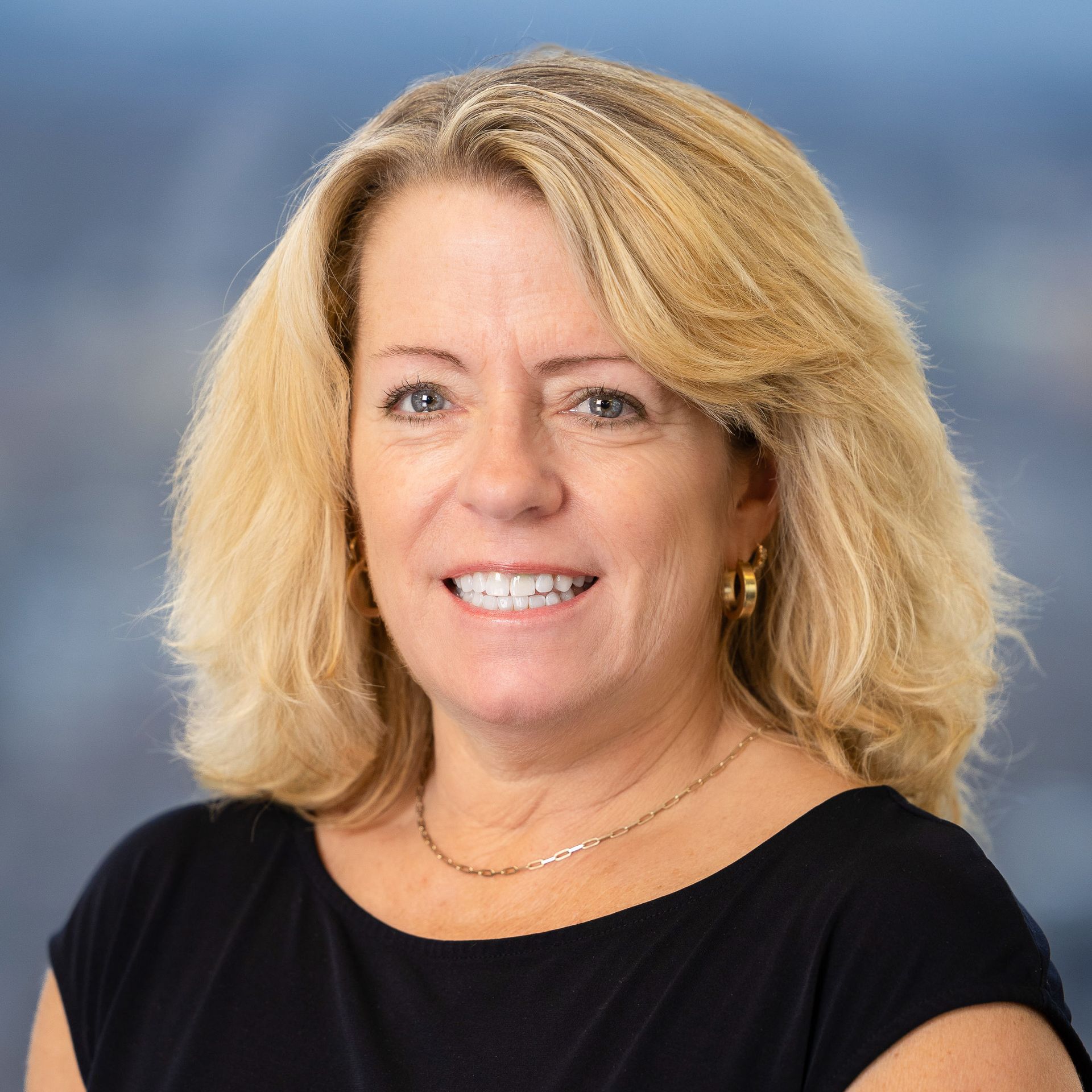 Blonde woman smiles, wearing a black top and gold jewelry, against a blurred background.