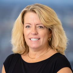 A portrait of Kelly Tubridy, Chief Executive Administrator of CONNSTEP,  smiling, wearing a black top and gold jewelry, against a blurred background.