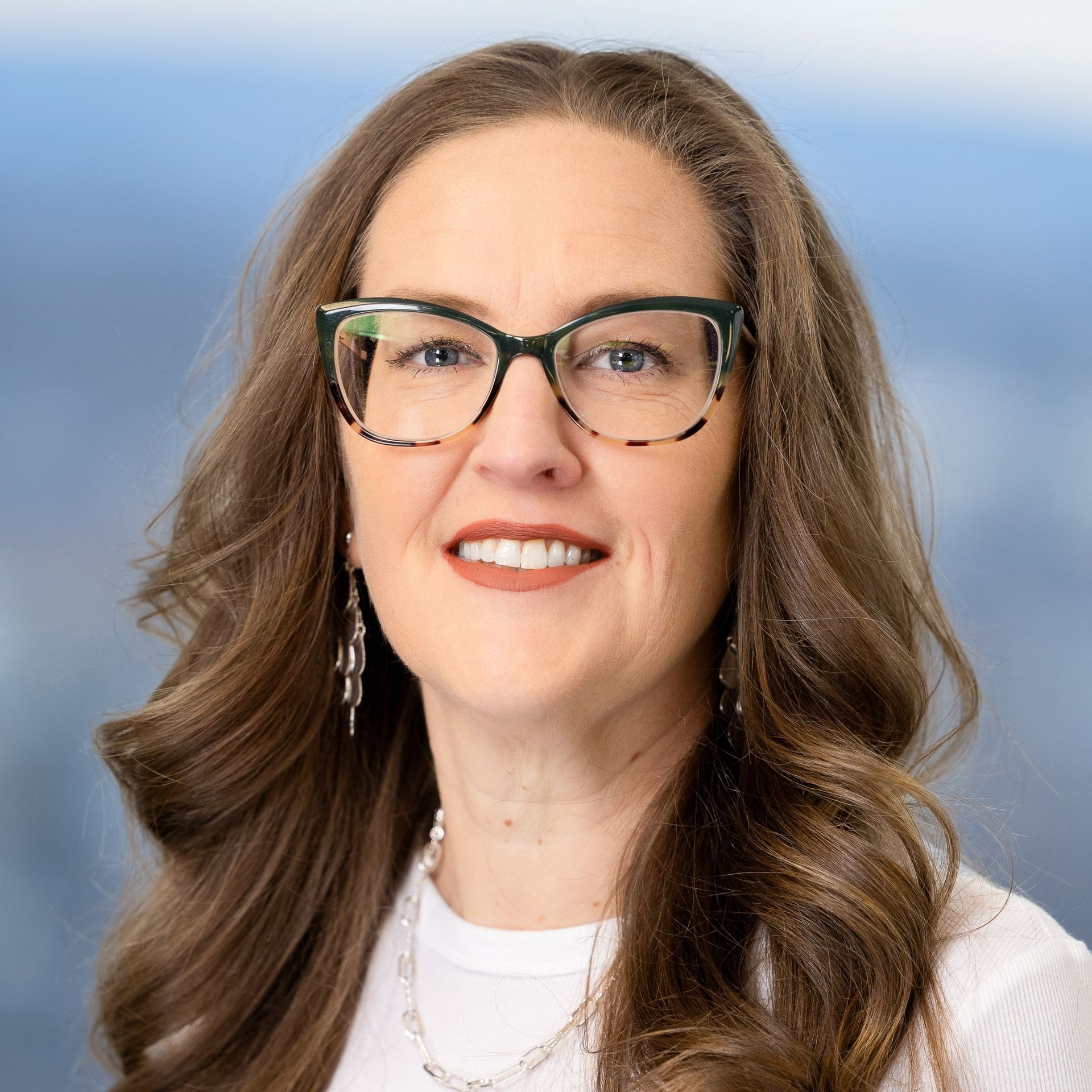 Woman with long brown hair, glasses, and a white top, smiling at the camera.