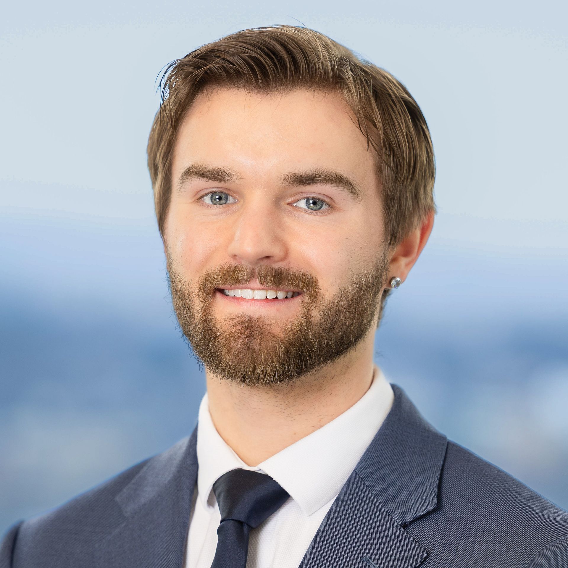 Man with light brown hair and beard, smiling, wearing a blue suit and tie, against a blurred background.