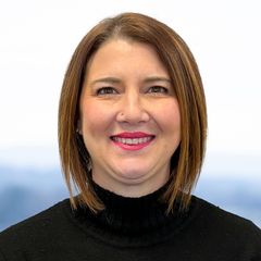 A portrait of Rachele Brown, Quality Management at CONNSTEP, with short brown hair, wearing a black turtleneck, smiling. Blurred, blue background.
