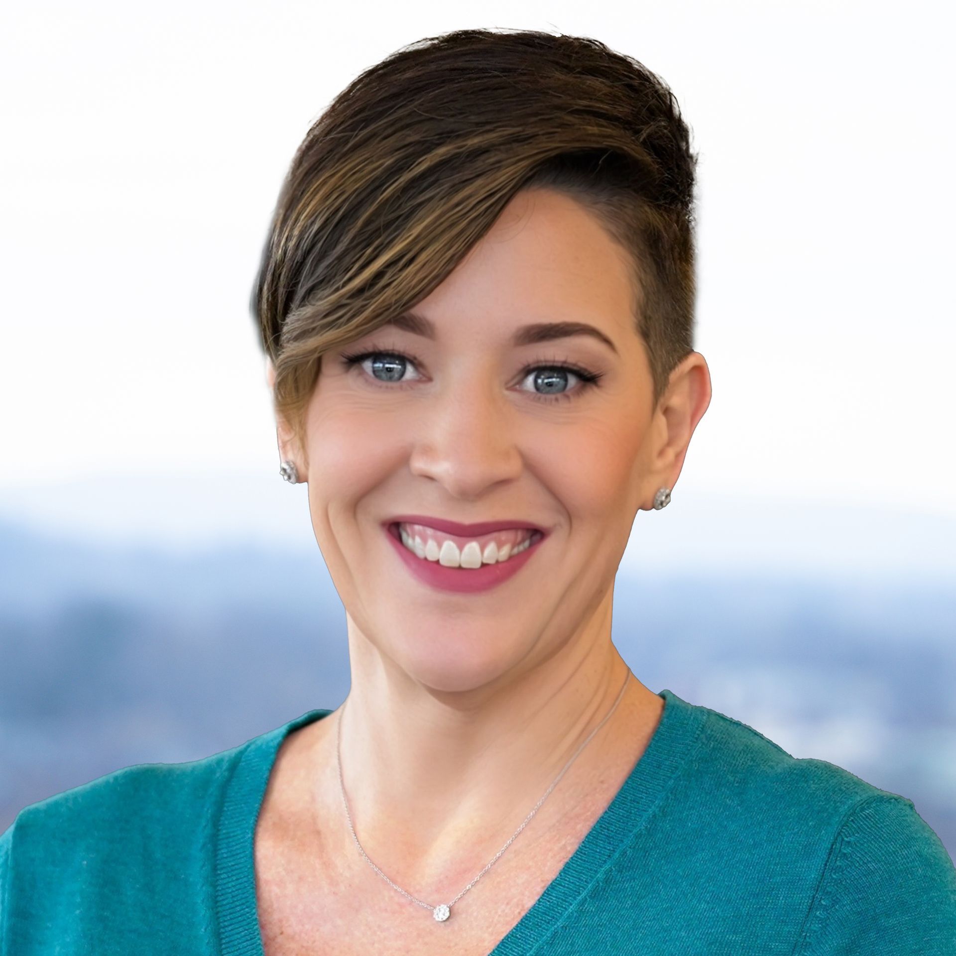 Woman with short hair, smiling. Wearing teal top and necklace. Blurred cityscape background.