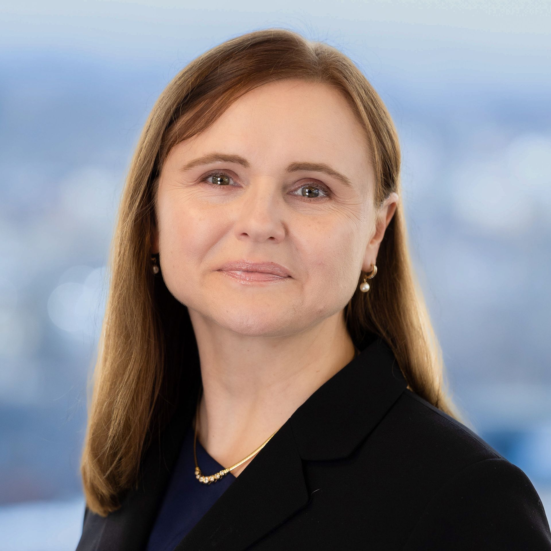 Woman in a black blazer smiles, looking at the viewer. Background: blurred cityscape.
