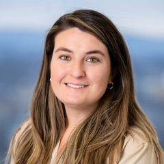 A portrait of Corinne McHenry, Business Development Advisor at CONNSTEP, with long, light brown hair smiling, wearing a beige shirt, with a blurred blue and white background.