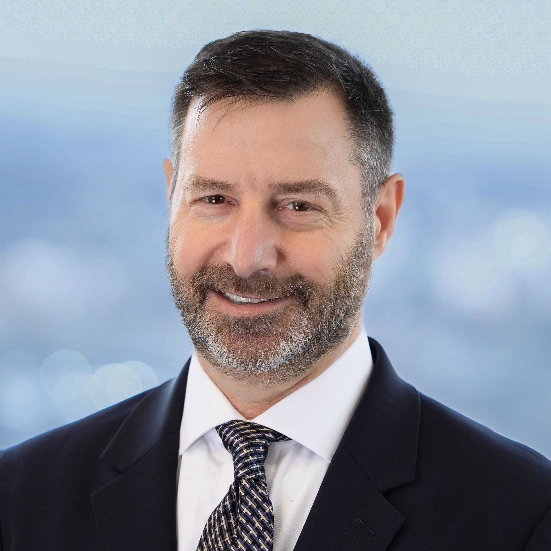 Man with a beard in a suit smiling at the camera, blurred city background.