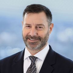 A portrait of Doug Narins, Business Development Advisor at CONNSTEP, with a beard in a suit smiling at the camera, blurred city background.