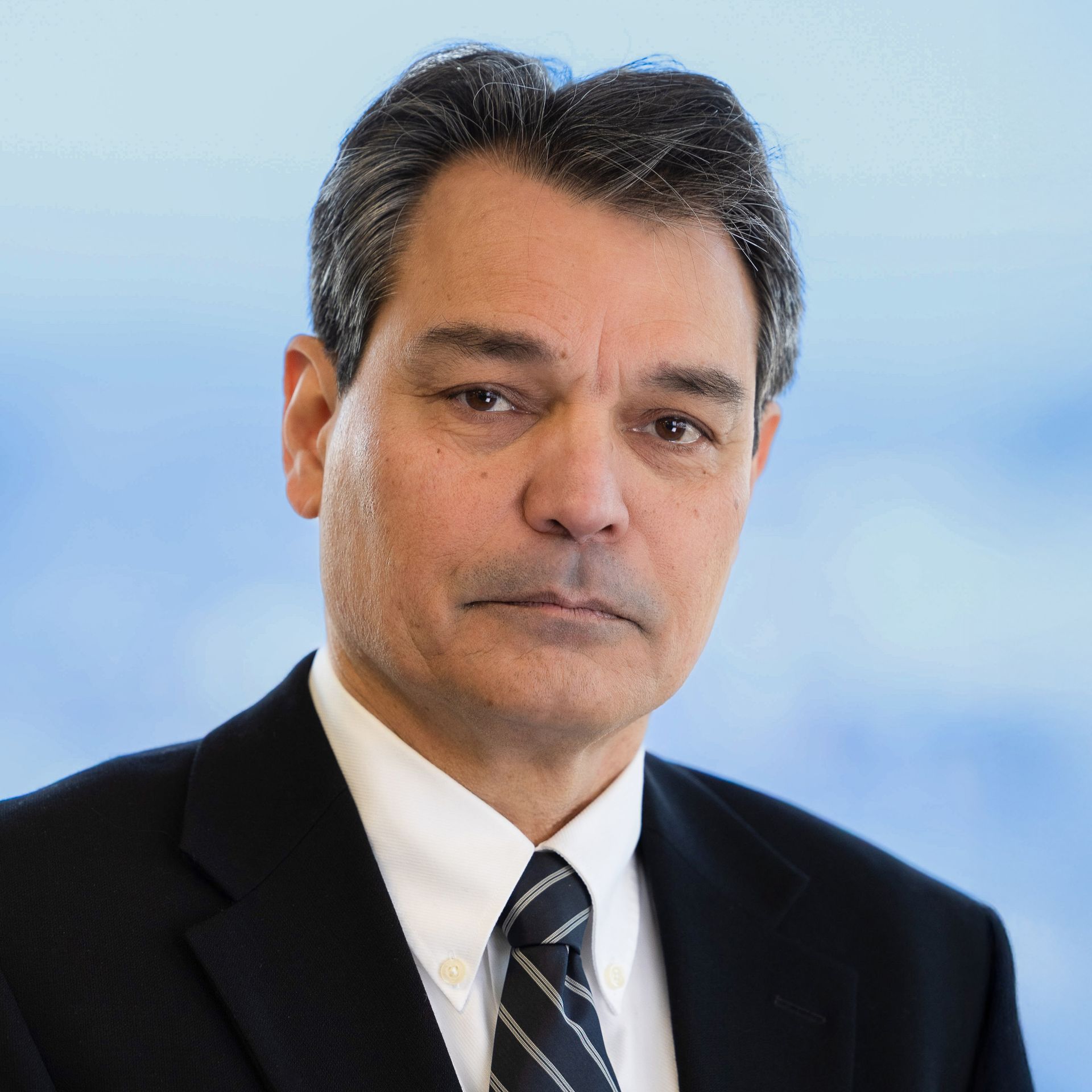 Man in a suit, looking at the camera with a neutral expression. Blurred blue background.