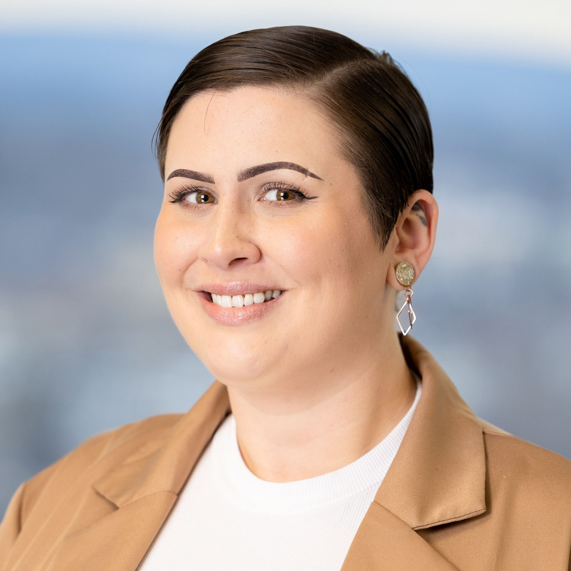 Woman with short dark hair, smiling, wearing a beige blazer and dangling earrings.