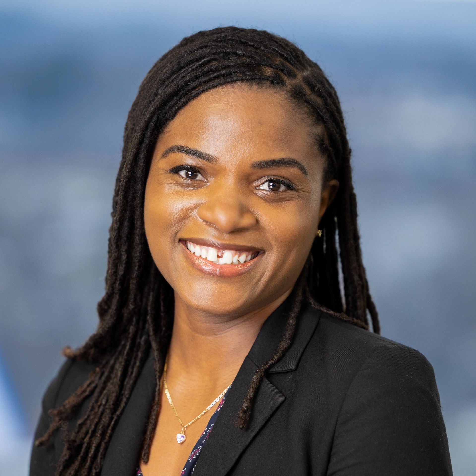 Woman with dark skin smiles, wearing a black blazer, against a blurred background.