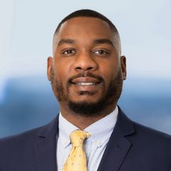 A portrait of Ricky Cotton, Business Services Associate at CONNSTEP, in navy suit, yellow tie, smiling. Blue and white blurred background.