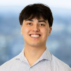 A portrait of Ryan Scott, CONNEX Supply Chain Database at CONNSTEP, smiling, in a patterned shirt, looking towards the camera, against a blurred blue background.
