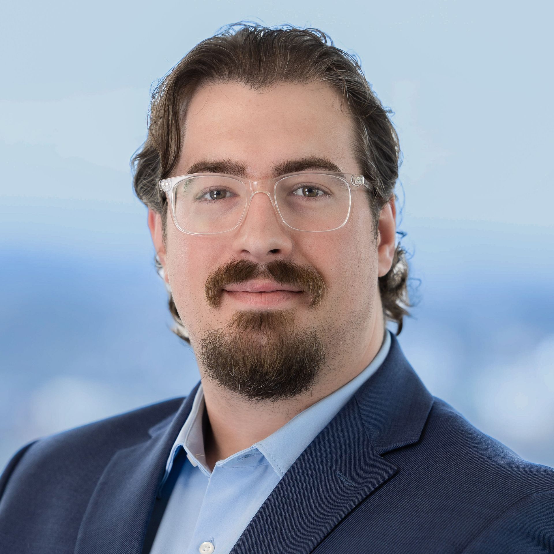 Man in glasses and blue suit smiles, facing the camera. Blurred blue background.