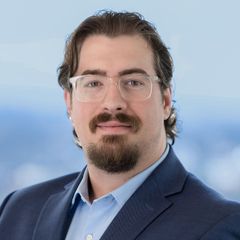 A portrait of Tyler Scott, Director, Marketing & Product Development at CONNSTEP, wearing glasses and blue suit smiling, facing the camera. Blurred blue background.
