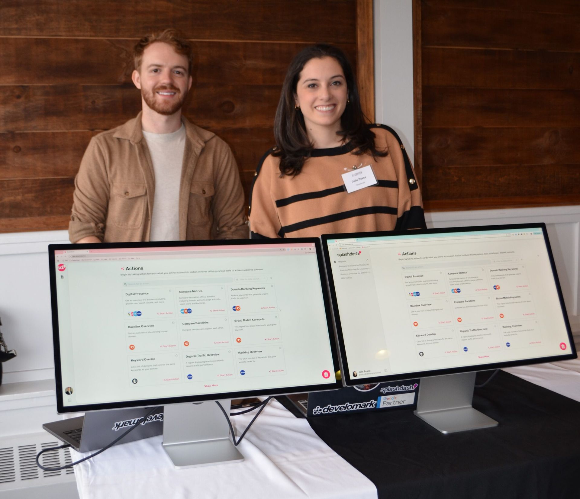 Two people stand behind two computer monitors displaying a website with icons. Monitors sit on a table with a black and white tablecloth.