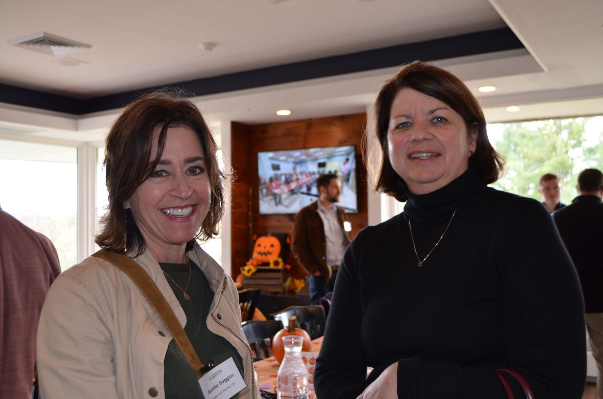 Two women smiling indoors, one in beige jacket, the other in black turtleneck, near a table and a TV.