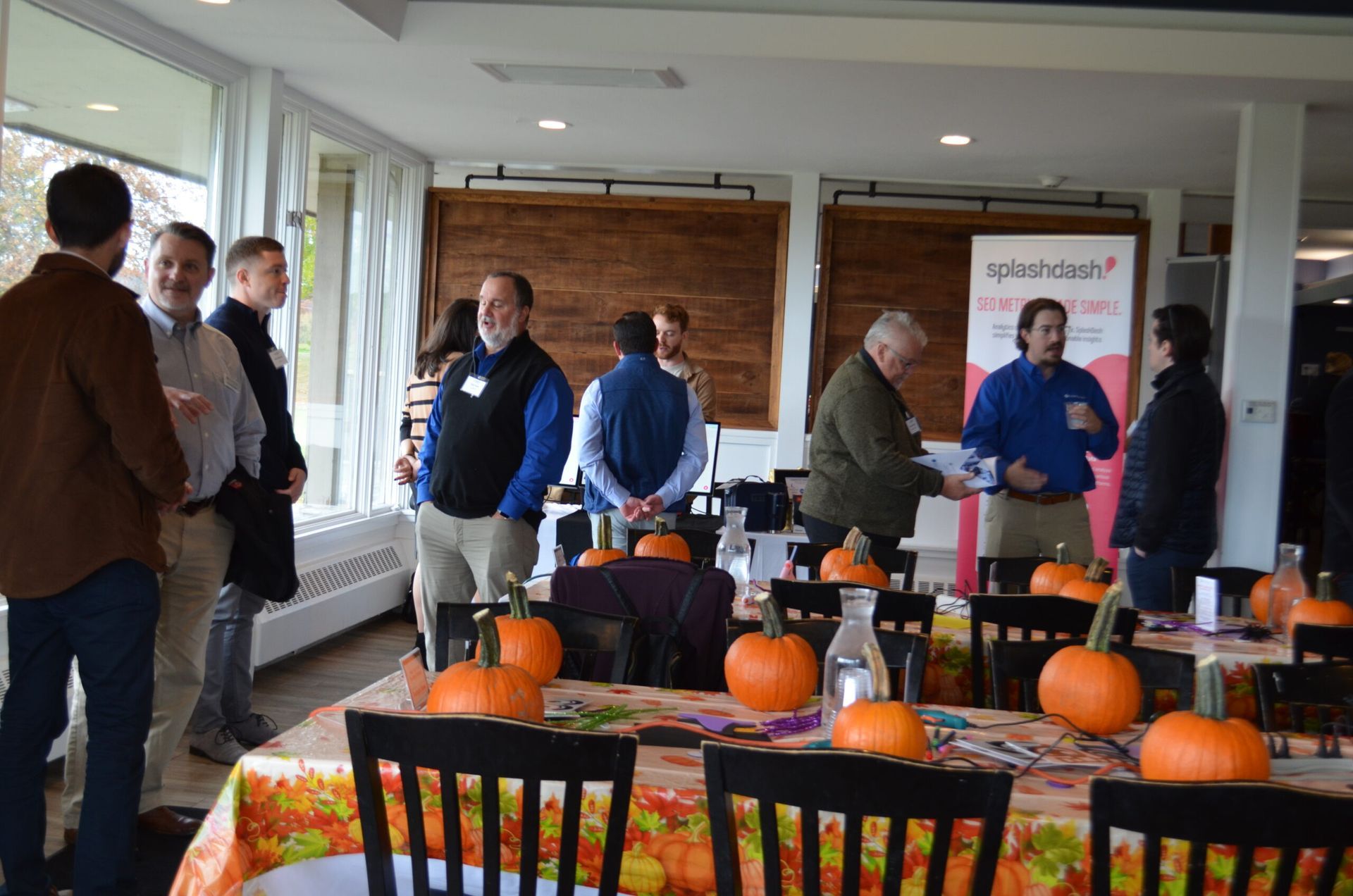 People at tables with pumpkins, socializing in a room with large windows, possibly a networking event.