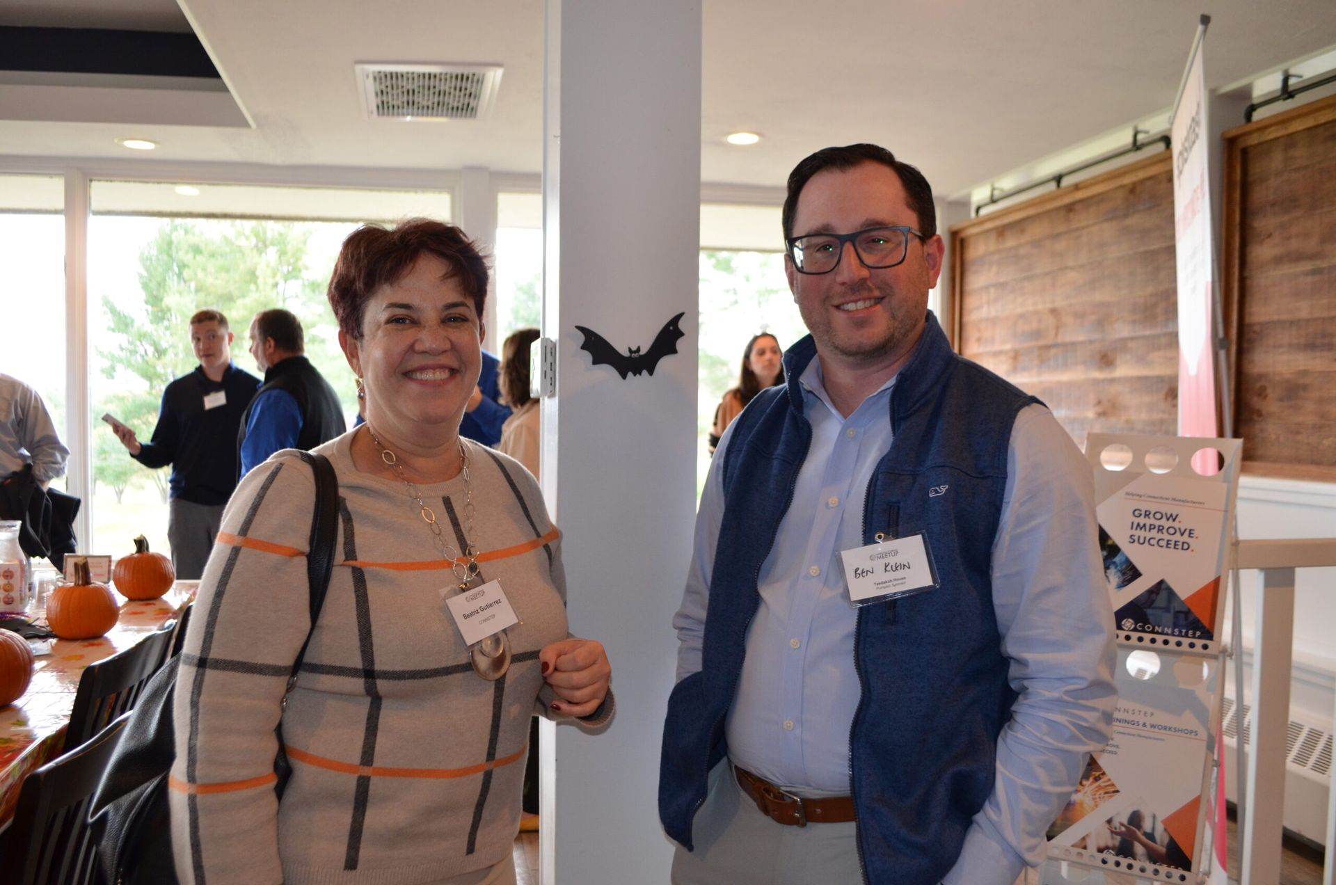 Woman and man smiling, standing by a pillar at an event. Pumpkins and brochures are in view.