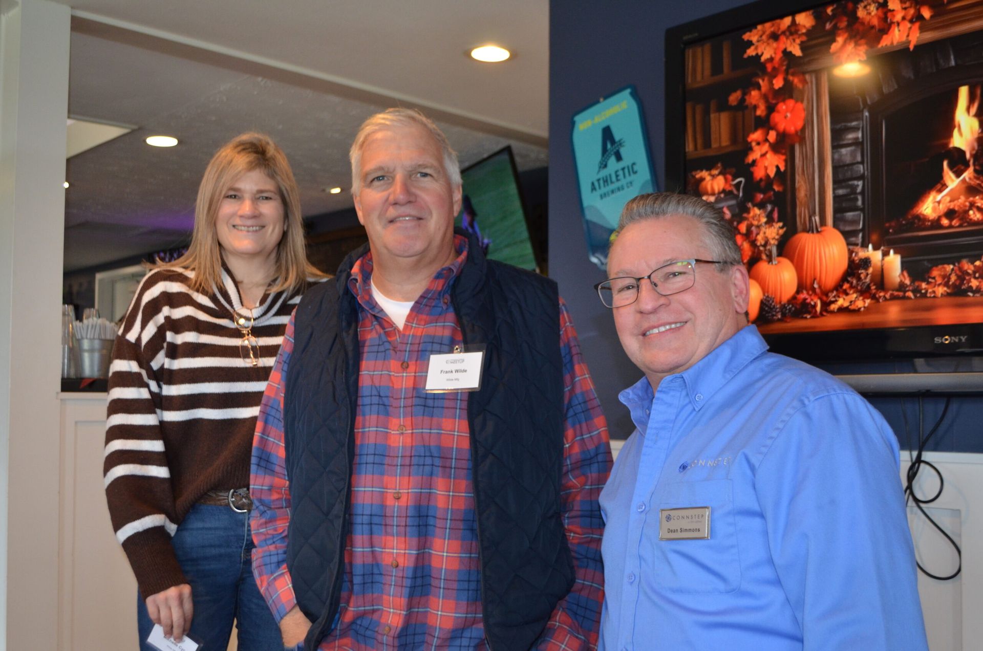 Three people smiling indoors, standing by a TV showing a fireplace scene.