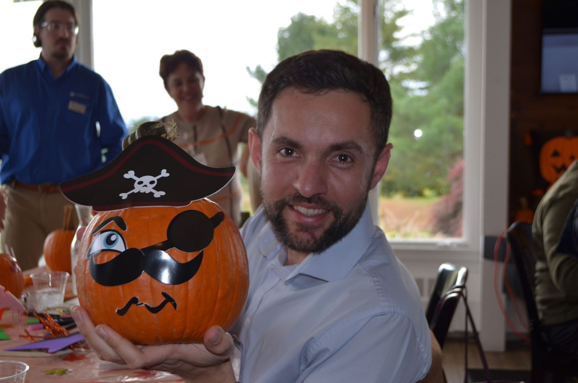 Man holding a decorated pumpkin with a pirate hat and winking eye; other people in the background.