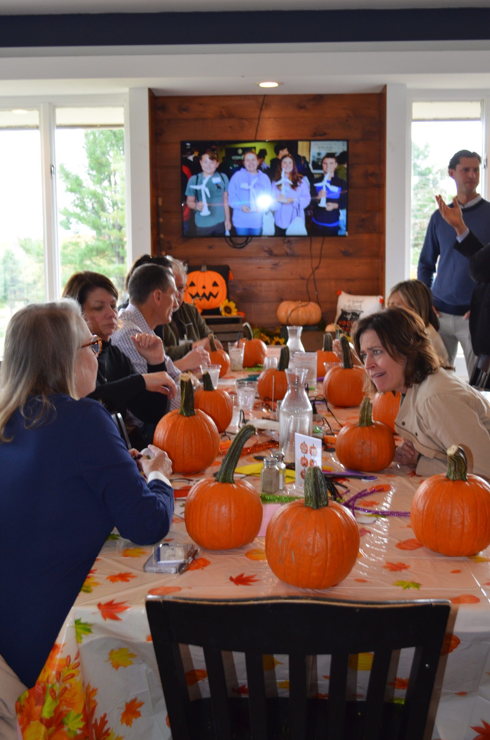 People at a table decorated with pumpkins, watching TV. Fall foliage tablecloth, bright and inviting.