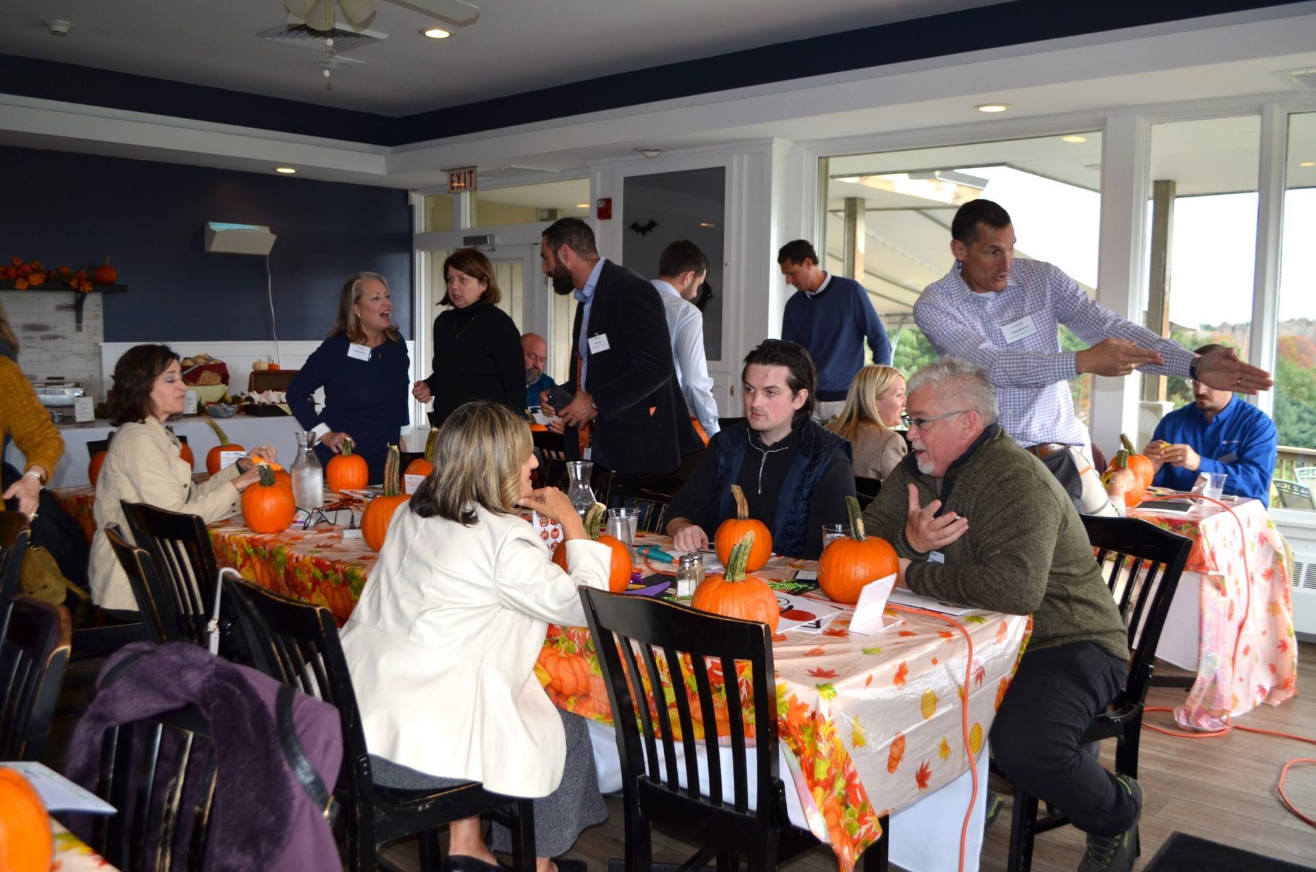 People at tables set with pumpkins, a fall-themed gathering. Some people are talking, gesturing, or standing.