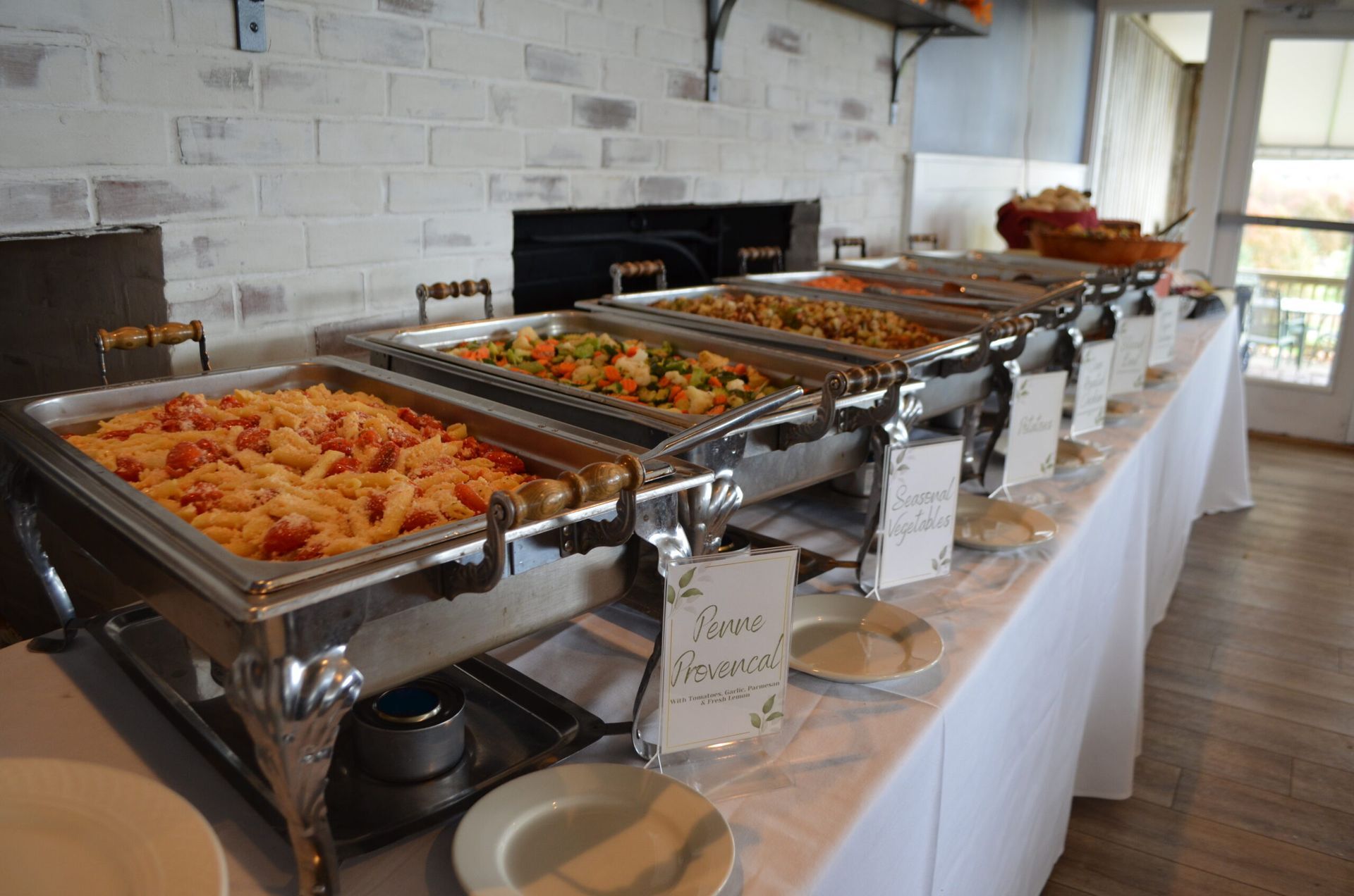 Buffet table with several chafing dishes of food, white tablecloth, brick wall background.