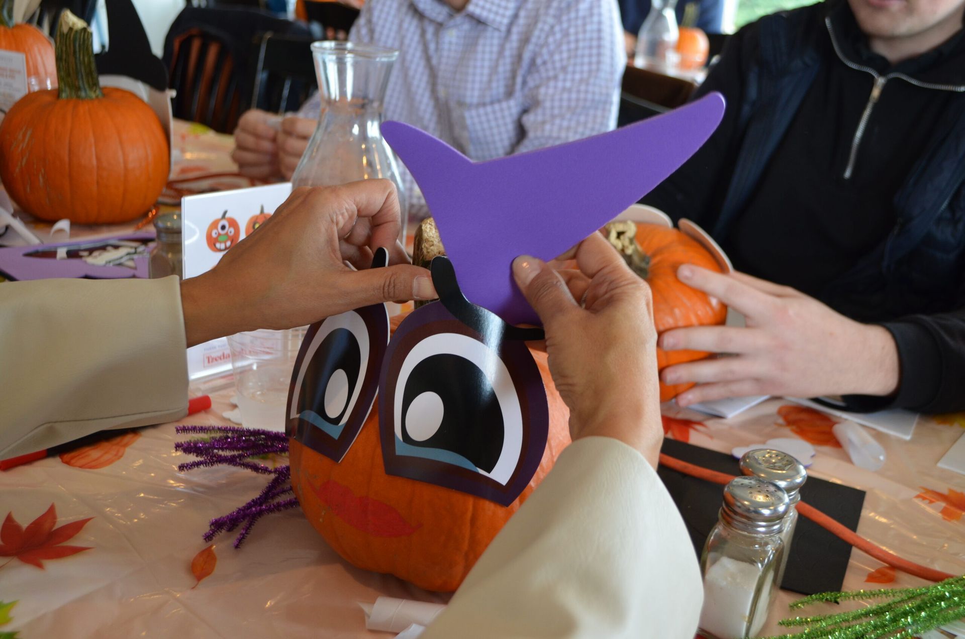Hands attach purple paper to a pumpkin decorated with large eyes and a beak.